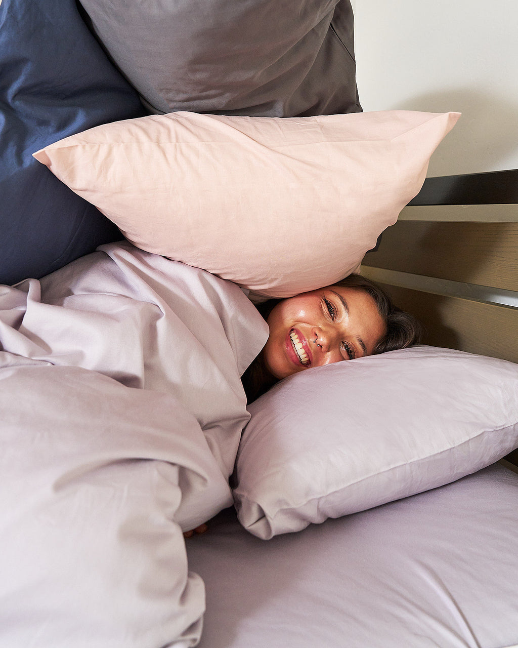 Twin XL Duvet and Pillowcases. Dorm Student laughing in bed on a lavender purple fitted sheet with side storage, wrapped in a l100% Cotton, 400 thread count avender purple duvet and her face in between fluffy pillows