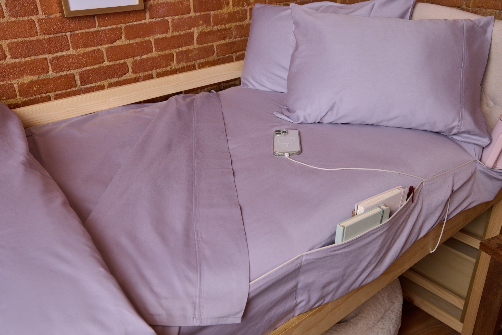 Bed with purple bedding and electronic devices on a wooden floor against a brick wall.